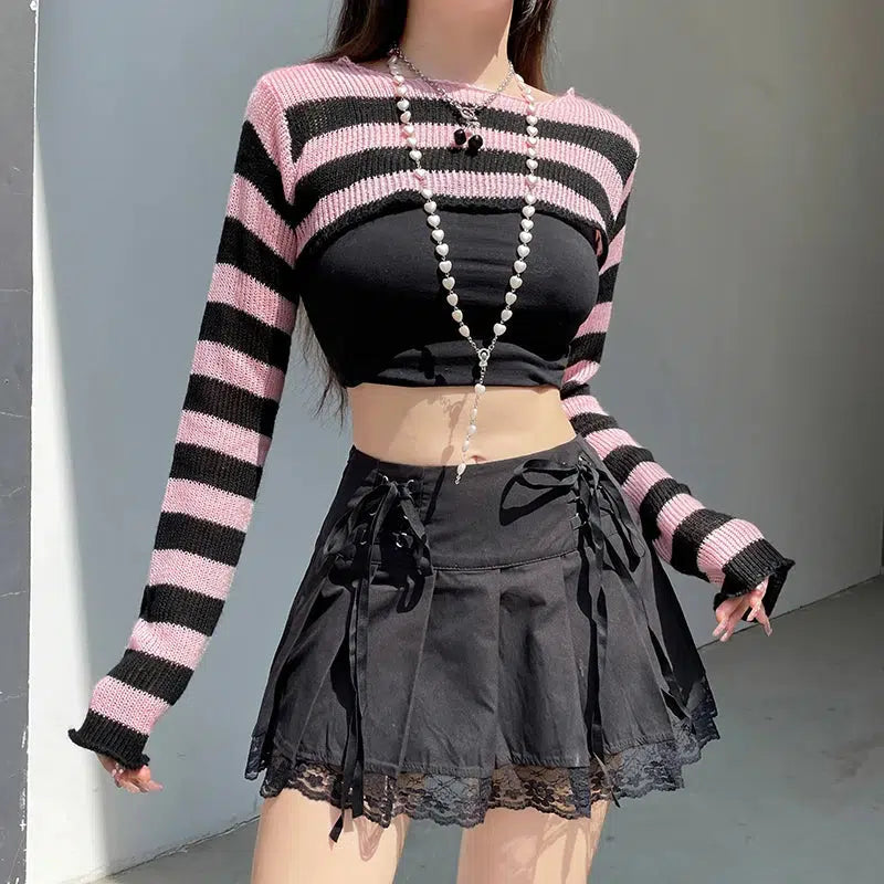 a girl is wearing a soft goth striped knitted shrug sweater in black and pink and she is also wearing a ribbon and lace detailed pleated skirt in black and a black top and a pearl necklace